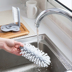 Hand holding a white and gray Kitchen Bottle Brush under running water in a stainless steel sink.